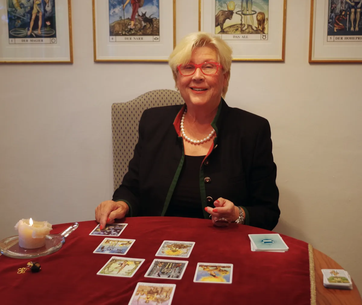 Eine Frau sitzt an einem Tisch mit rotem Tischtuch, einer Kerze und Tarotkarten. 